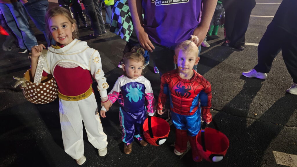 Three children in Halloween costumes at a Trunk or Treat event in St. Charles County with a parent wearing a Doggy Doods shirt.